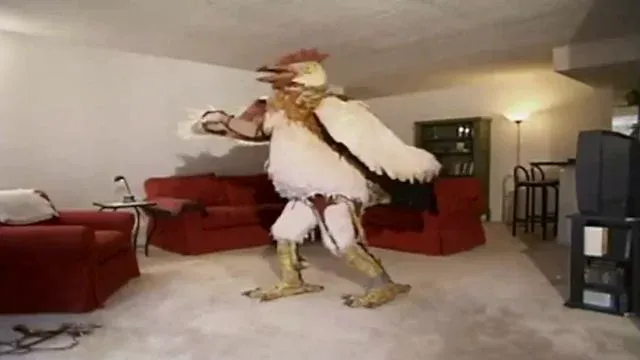Poorly compressed screenshot of a man in a chicken costume, with the arms sticking out of the wings and garters holding up the feet. He's in a small room, windowless, with a low ceiling, beige carpet, red sofa, Ikea lamp and old CRT TV on a stand. It's bizarre.